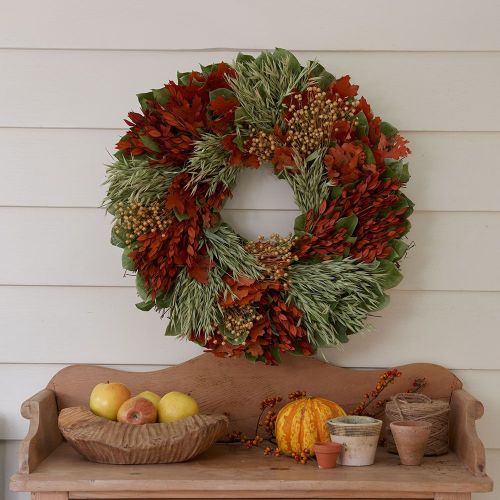 A colorful autumn wreath made of leaves and berries hangs on a wall above a wooden bench with apples and decorative gourds.