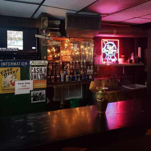 A dimly lit bar with a neon sign, shelves of bottles, and a cash-only sign on the counter.
