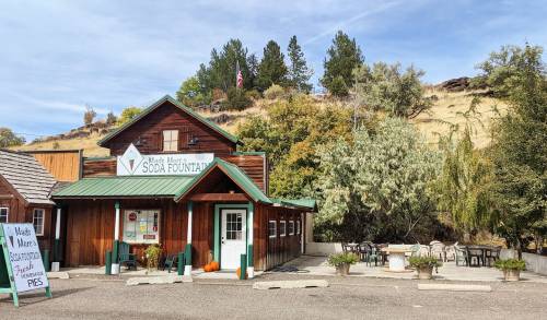 A rustic soda fountain shop with a wooden exterior, surrounded by trees and a small outdoor seating area.