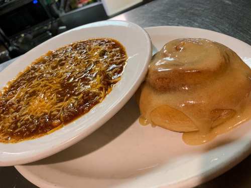A plate with chili topped with cheese and a cinnamon roll drizzled with icing.