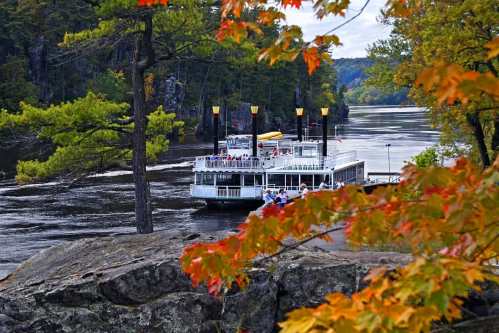 A riverboat with passengers sails along a scenic river, surrounded by autumn foliage and rocky shores.