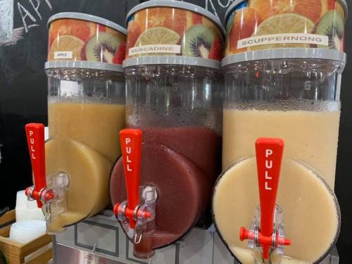 Three beverage dispensers filled with apple, muscadine, and scuppernong slushies, each with a red pull handle.