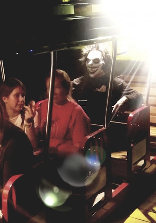 A spooky figure with a skull mask sits behind two people on a ride, illuminated by bright lights.