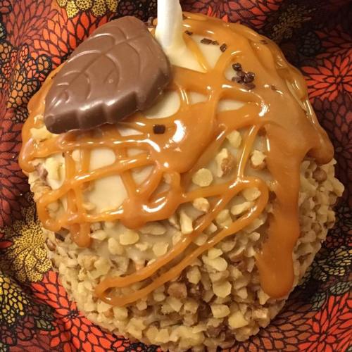 A caramel-covered apple coated in chopped nuts, topped with a chocolate piece and drizzled with caramel.