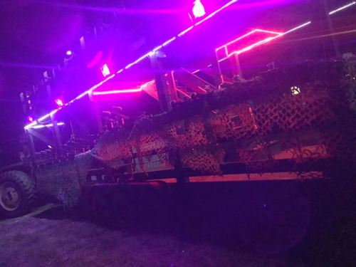 A trailer covered in camouflage netting, illuminated by vibrant purple and pink lights at night.