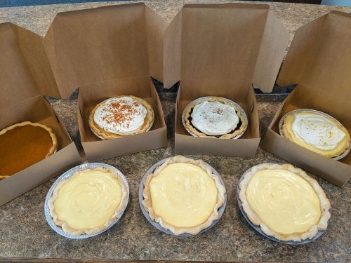 A variety of pies in cardboard boxes, including pumpkin, cream, and chocolate, displayed on a countertop.