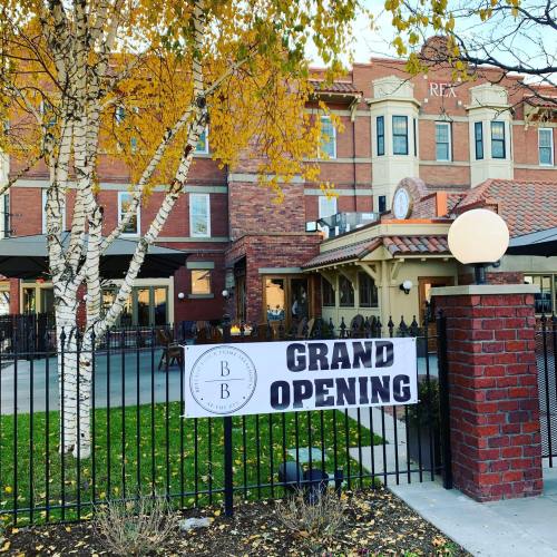 A brick building with a "Grand Opening" banner in front, surrounded by autumn trees and outdoor seating.