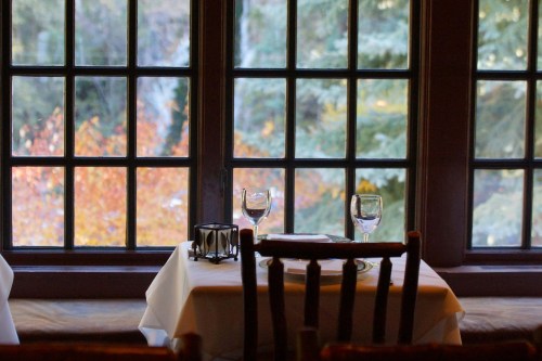 A cozy restaurant table set for two, with wine glasses, near a large window showcasing autumn foliage outside.