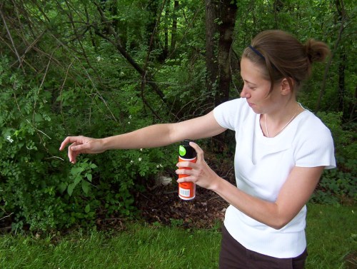 A woman in a white shirt sprays insect repellent on her arm while standing in a lush green outdoor setting.