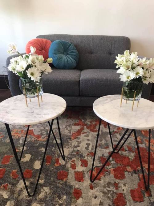A gray couch with colorful cushions between two marble-top side tables holding flower arrangements, on a patterned rug.