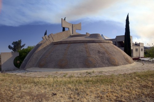 A unique, dome-shaped building with a cross on top, surrounded by trees and a clear blue sky.
