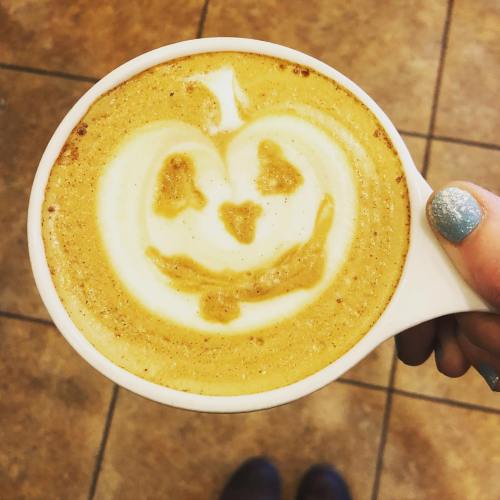 A hand holding a latte with a pumpkin face art design on the frothy surface.