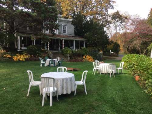 A garden with white tables and chairs set up on green grass, surrounded by trees and a house in the background.