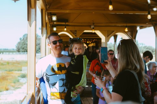 A man in a Batman shirt holds a smiling girl in a cape, while a woman takes a photo of them on a sunny day.