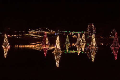 Colorful Christmas lights illuminate trees and a house, reflecting beautifully on a calm pond at night.