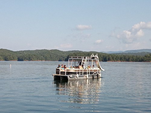 A pontoon boat with several people is cruising on a calm lake surrounded by green hills and a clear blue sky.