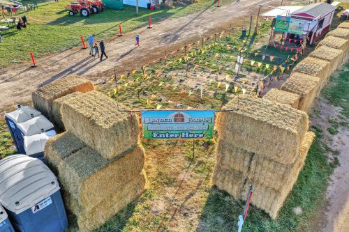 Aerial view of a farm entrance marked by hay bales and a sign reading "Enter Here," with people walking in the background.
