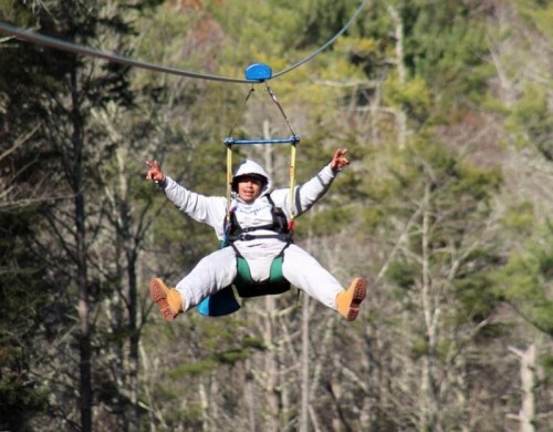 A person in a gray outfit zip-lining through a forest, smiling and making a peace sign with their hands.