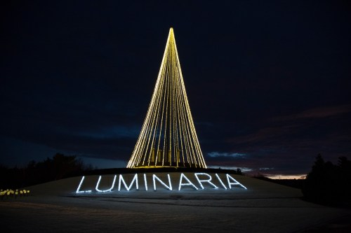 A tall, illuminated Christmas tree with golden lights stands on a hill, with the word "LUMINARIA" displayed in bright letters below.