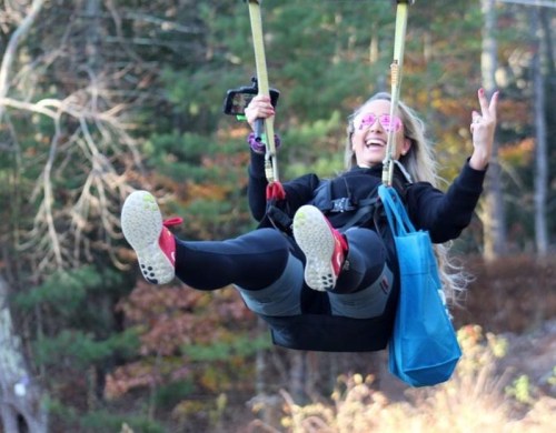 A woman with long hair swings joyfully, wearing pink sunglasses and a black outfit, surrounded by trees in autumn.