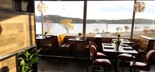 Cozy restaurant interior with wooden booths, tables set for dining, and a scenic view of a lake and trees outside.