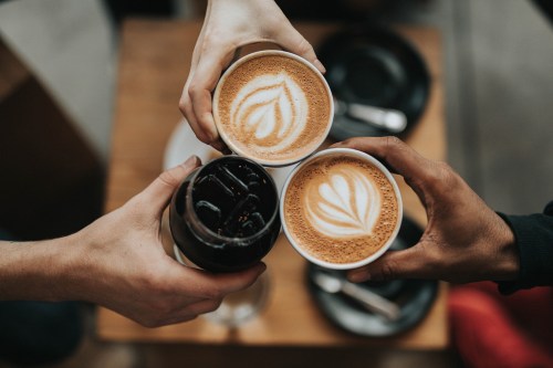 Three hands hold coffee cups with latte art, clinking together over a wooden table.