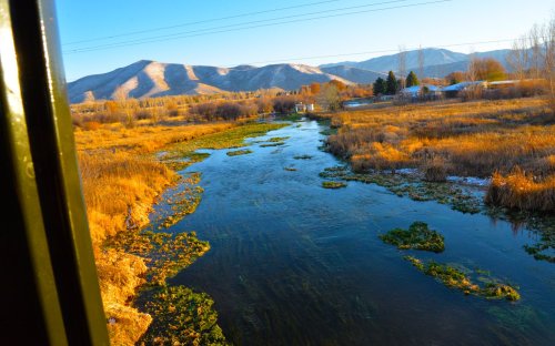 A serene river flows through golden grasslands, with mountains in the background under a clear blue sky.