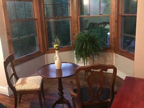 Cozy corner with a small table, two chairs, a vase of flowers, and a potted fern by large windows.