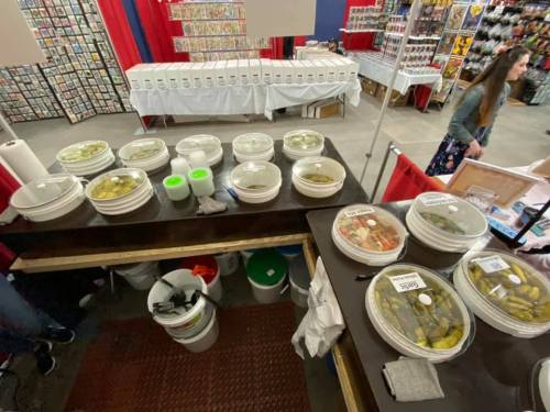 A vendor's table with various containers of pickles and snacks at a market or fair, with people browsing in the background.