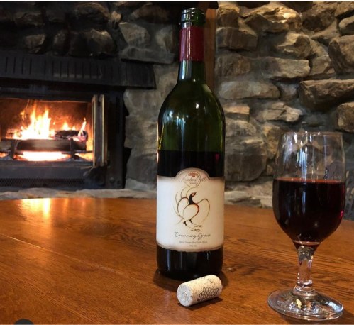 A bottle of red wine and a glass sit on a wooden table near a cozy stone fireplace. A cork lies beside the bottle.
