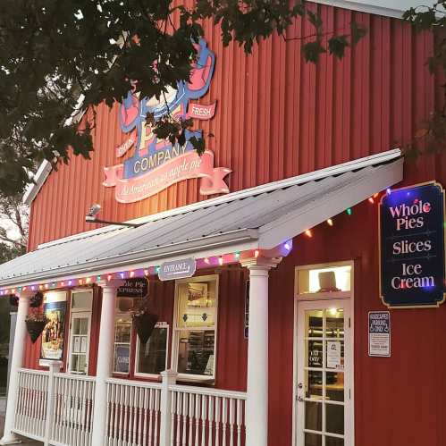 Red wooden building with festive lights, featuring signs for whole pies, slices, and ice cream. Cozy, inviting atmosphere.