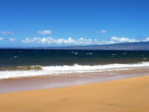 A serene beach scene with golden sand, gentle waves, and a clear blue sky dotted with clouds.