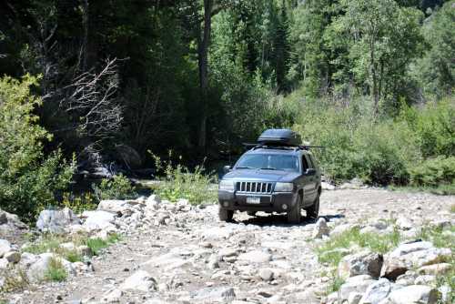 A black SUV parked on a rocky path near a river, surrounded by lush green trees and foliage.