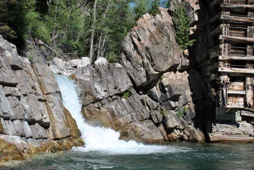 A waterfall cascades over rocky terrain, surrounded by lush greenery and a wooden structure nearby.