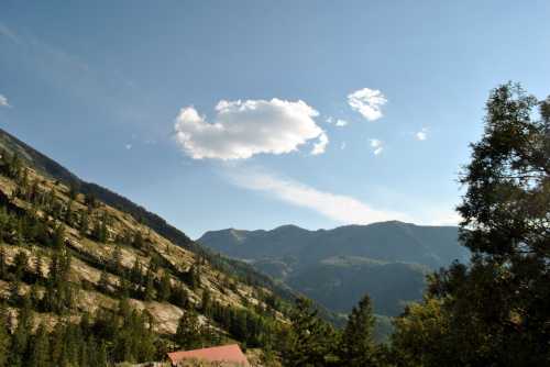 A scenic view of mountains under a clear blue sky with a few clouds and lush greenery in the foreground.