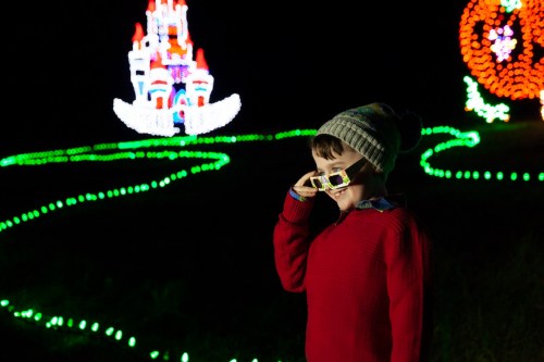A child in a red sweater and beanie poses with sunglasses, surrounded by colorful holiday lights at night.
