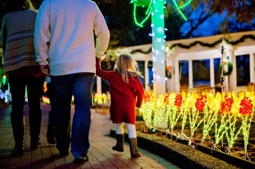 A child in a red coat holds an adult's hand while walking past colorful holiday lights in a festive setting.