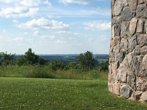 A stone wall stands in the foreground, overlooking a lush green landscape and blue sky with scattered clouds.