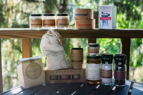 A variety of jars and packages of J.Q. Dickinson's gourmet salts and sauces displayed on a wooden surface outdoors.
