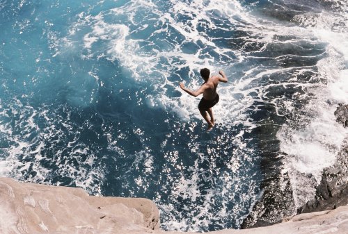A person jumps off a rocky ledge into vibrant blue ocean waters below. Waves crash around the rocks.