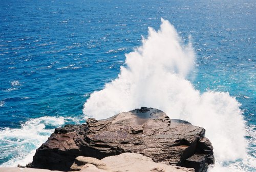 A rocky shoreline with waves crashing and spraying water against the vibrant blue ocean.