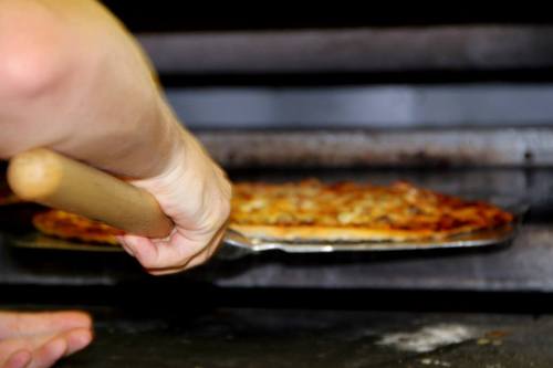 A hand using a pizza peel to remove a freshly baked pizza from an oven.