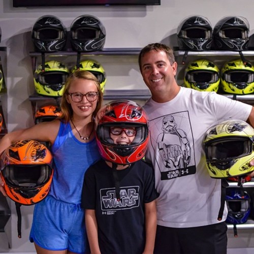 A man and two children hold motorcycle helmets in front of a wall displaying various helmets.