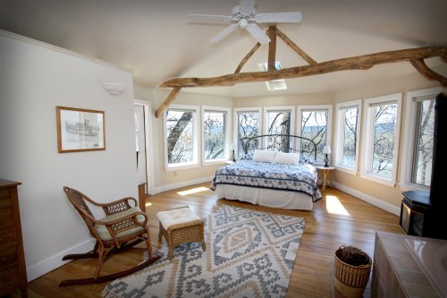 Cozy bedroom with large windows, a bed, rocking chair, and wooden accents, overlooking a serene outdoor view.