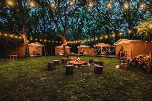 A cozy campsite with tents, a campfire, and string lights illuminating a grassy area surrounded by trees.