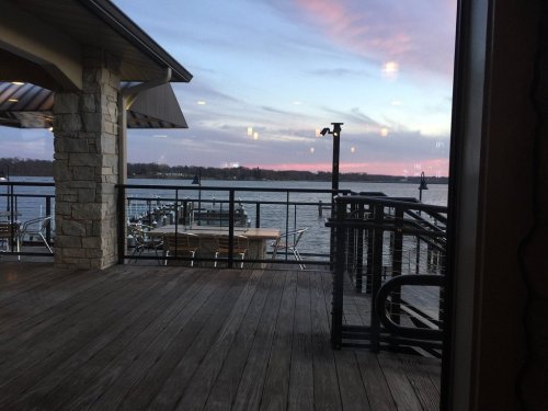 View of a serene lake at sunset from a wooden deck, with tables and chairs visible in the foreground.