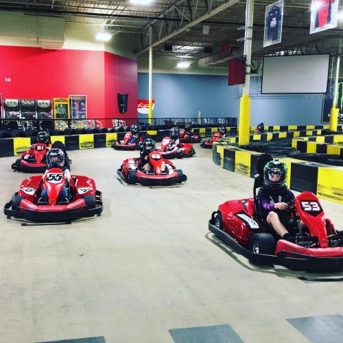 A go-kart track with several red go-karts racing, featuring drivers in helmets and a colorful indoor setting.