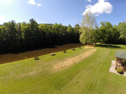 A serene landscape featuring a grassy area with benches, surrounded by trees and a calm water body under a blue sky.
