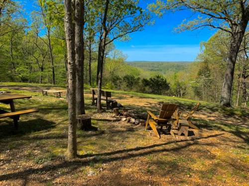 A serene outdoor scene with wooden chairs, a picnic table, and a fire pit, surrounded by trees and a distant view.