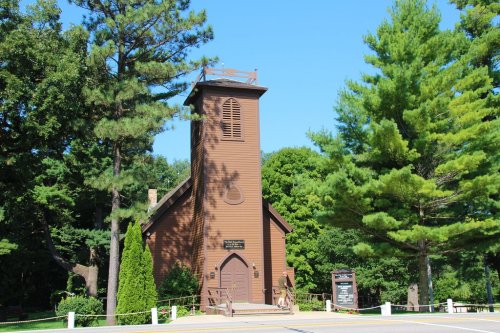 A brown wooden church with a tall steeple, surrounded by green trees and a clear blue sky.